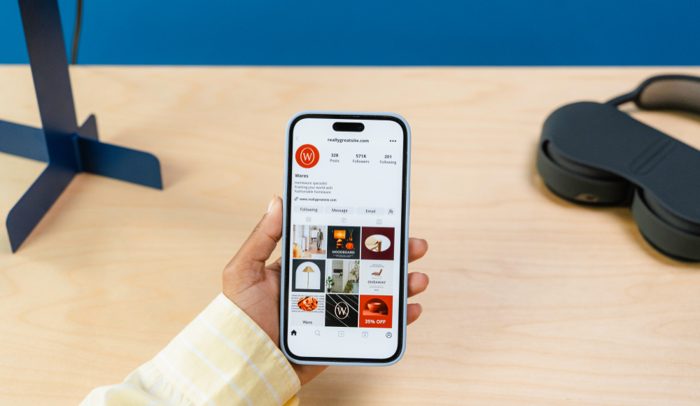 Person holding a smartphone displaying an Instagram profile with product images and a promotional discount offer, with a blue background and headphones nearby.