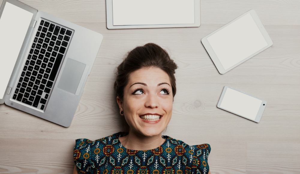 Smiling woman surrounded by a laptop, tablets, and smartphones, representing the multi-device access to Google Ads for business marketing.